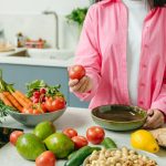 Woman in pink overshirt holds tomato in kitchen with fresh fruits and veggies.