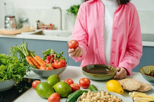 Woman in pink overshirt holds tomato in kitchen with fresh fruits and veggies.